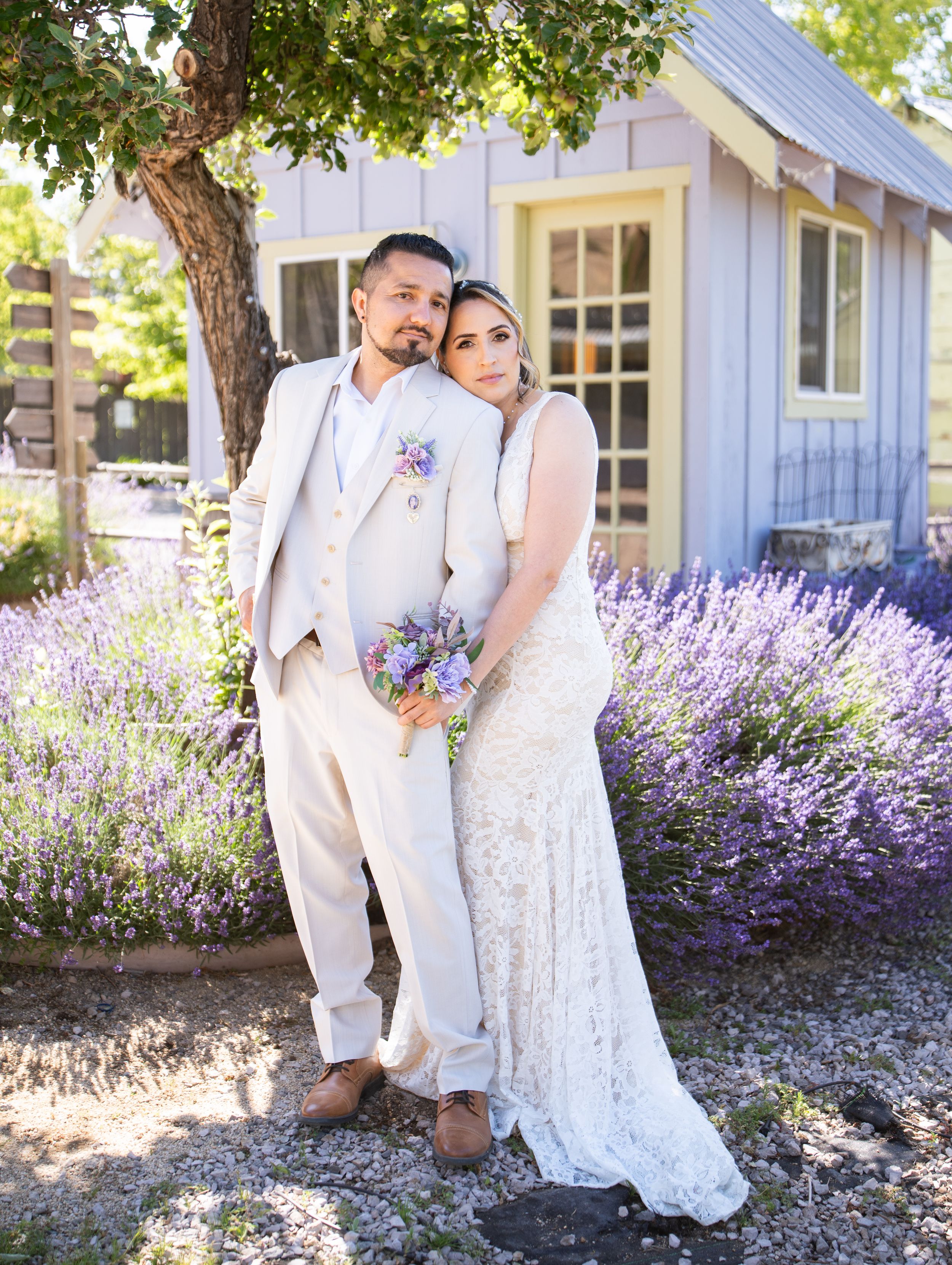 A wedding couple stands in front of a charming white cottage surrounded by blooming lavender flowers.