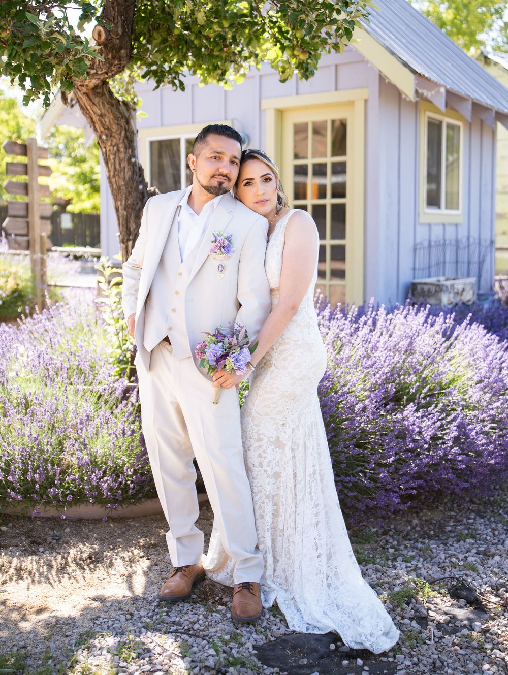 A wedding couple stands in front of a charming white cottage surrounded by blooming lavender flowers.
