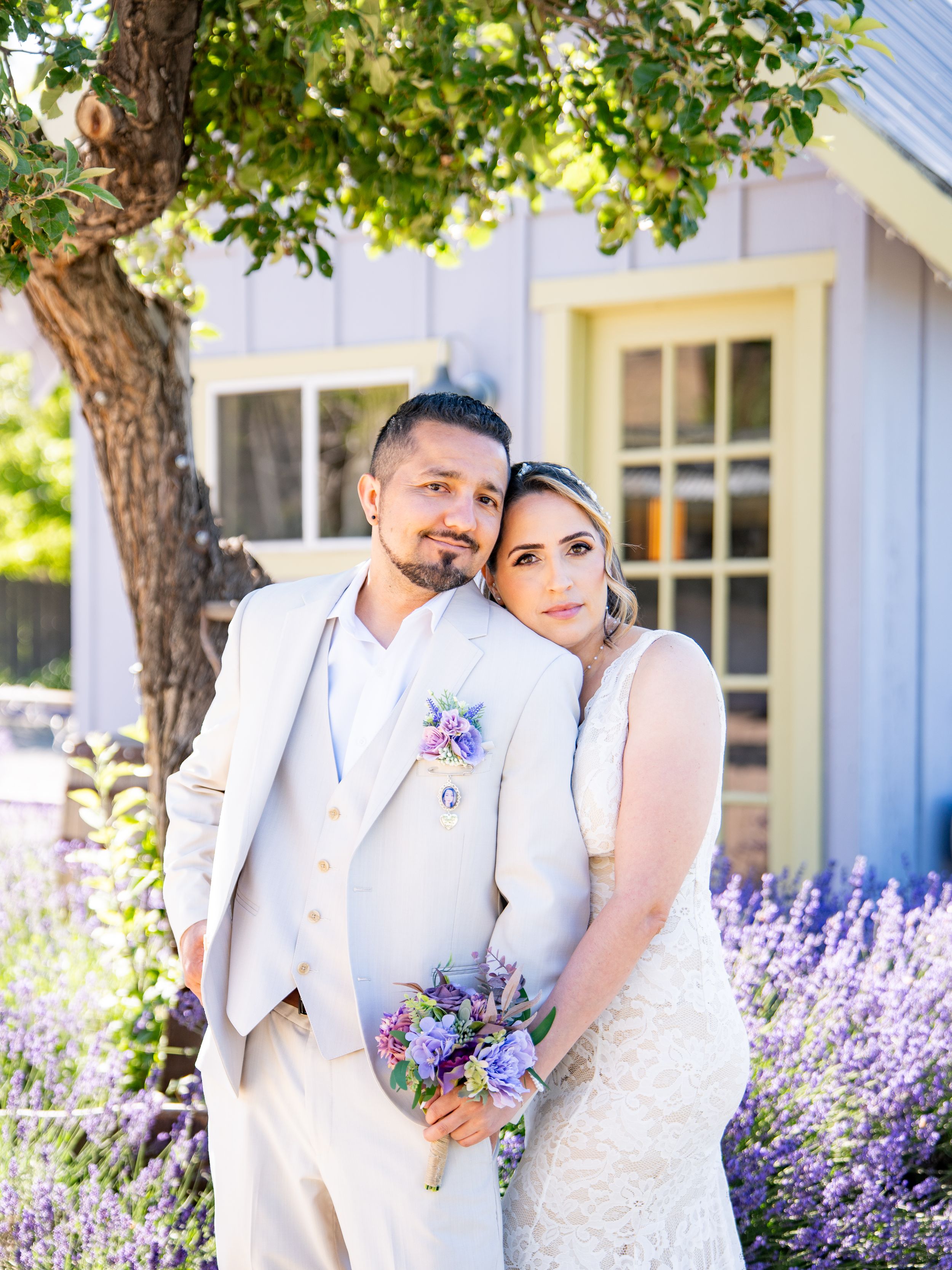 Wedding couple poses in front of a rustic white building with blooming purple lavender flowers and lush greenery.