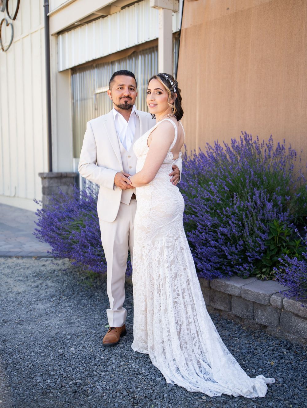 A couple poses together in formal wedding attire near blooming purple lavender bushes by a rustic building wall.