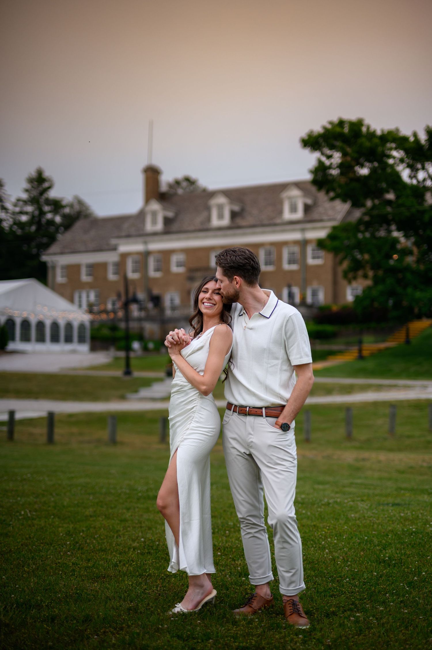 A joyful couple dances together on a grassy lawn with beautiful colonial buildings visible in the background.