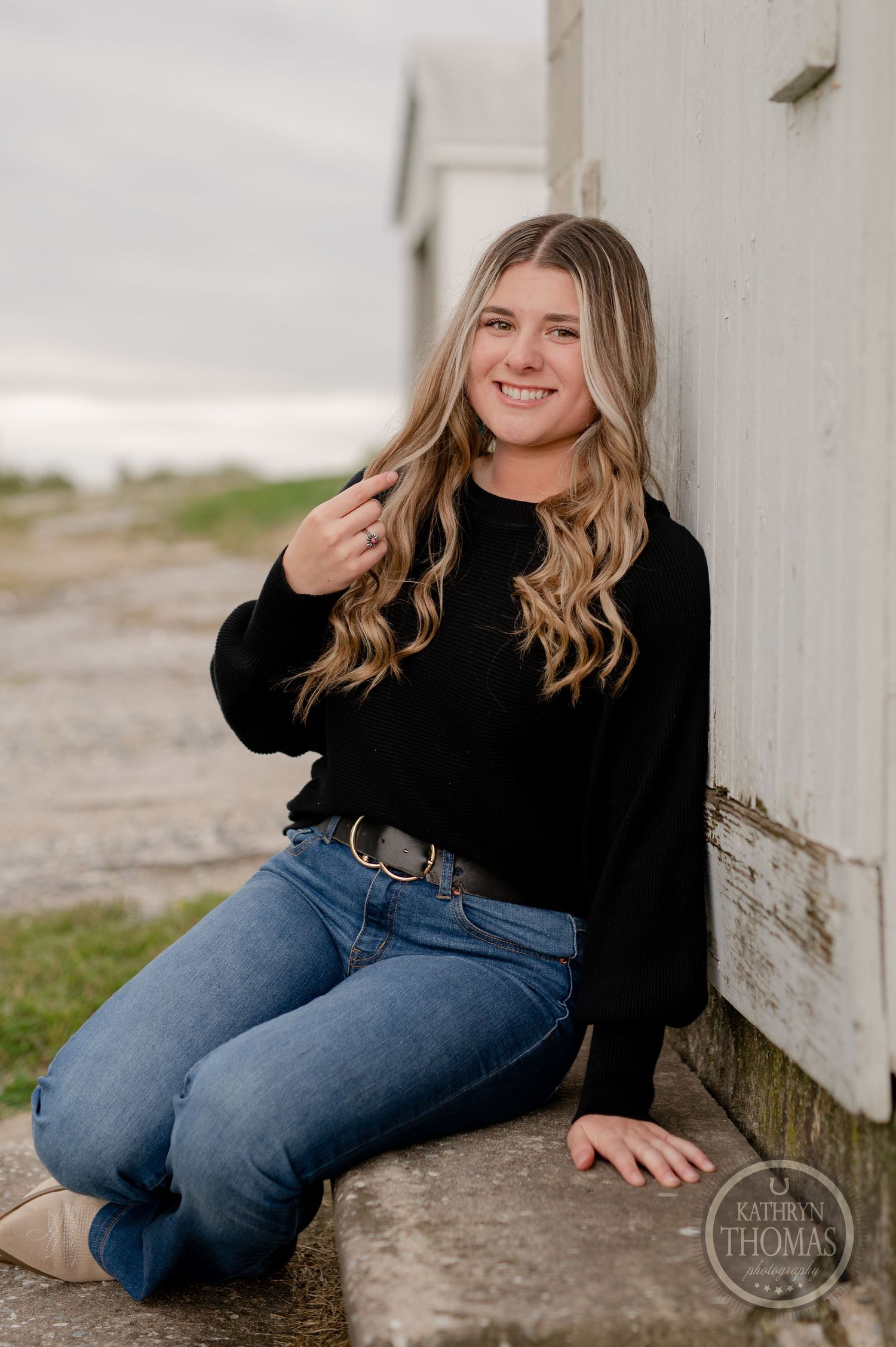Casual portrait of someone sitting by white barn wall wearing black sweater and jeans.