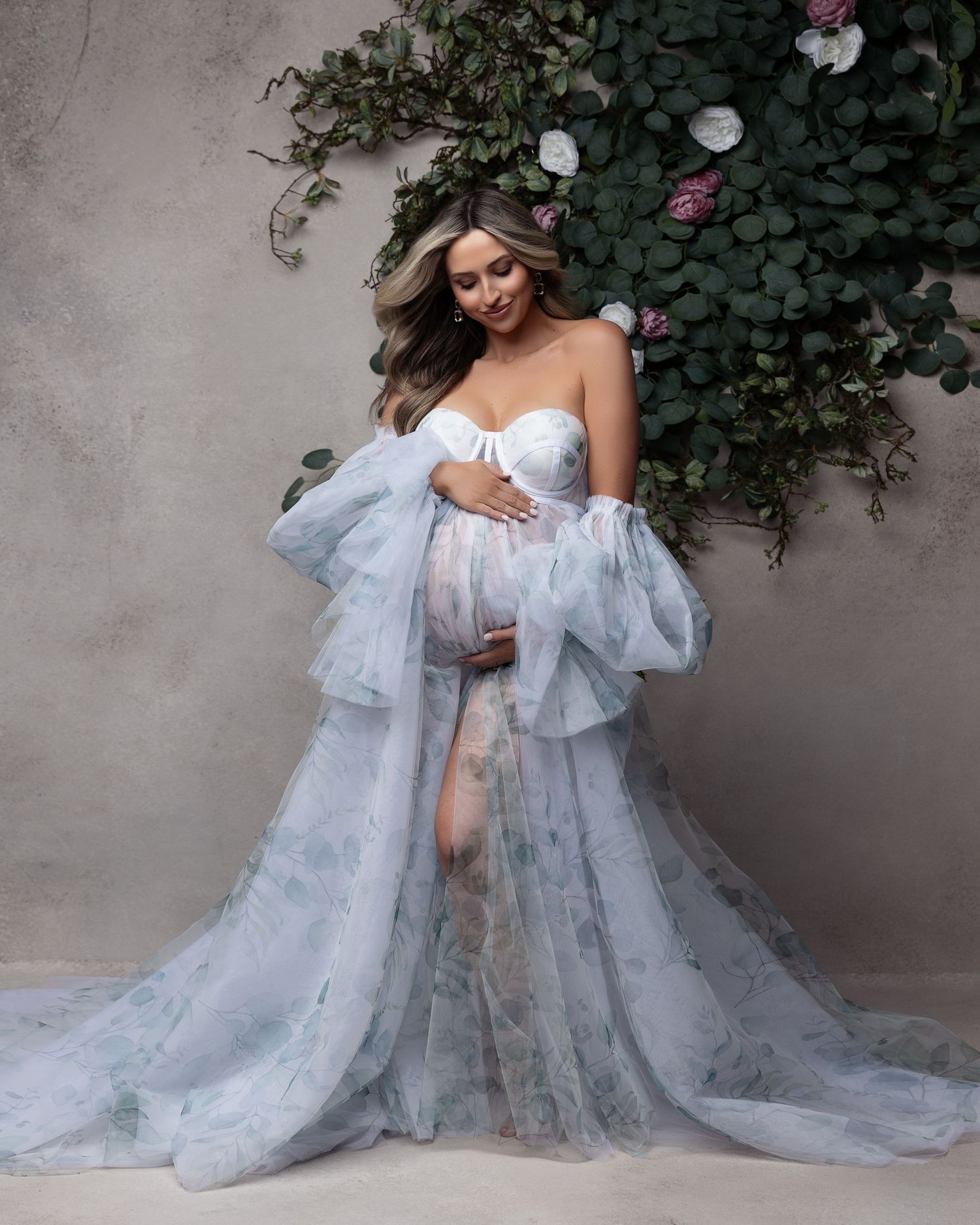 A dreamy maternity photoshoot features an ethereal light blue tulle gown against a garden wall with delicate flowers.