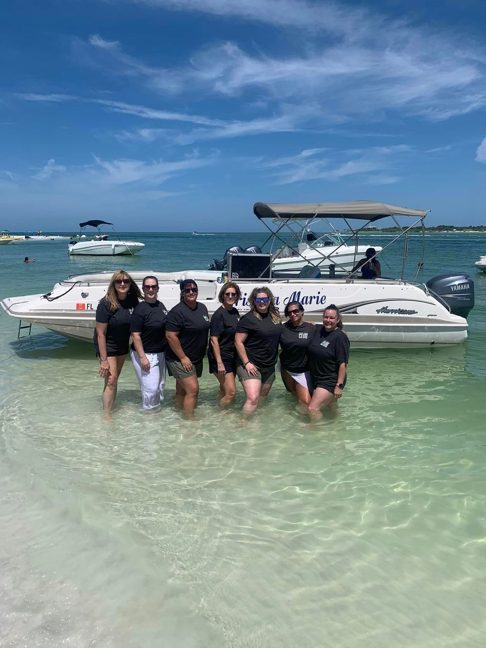 Group of women in black shirts posing in shallow turquoise water beside a white speedboat on a sunny beach day.