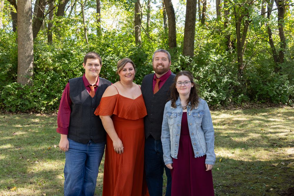 Family of four posing outdoors in semi-formal attire, smiling together in a lush green wooded setting.
