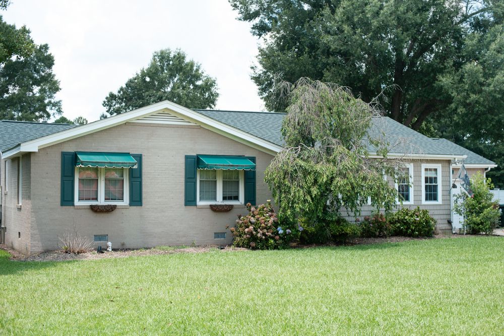 Small beige ranch-style house with teal awnings and shutters sits on a green lawn surrounded by mature trees.