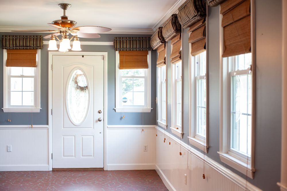 Bright sunroom with bamboo shades features white wainscoting, gray walls, and an elegant chandelier.