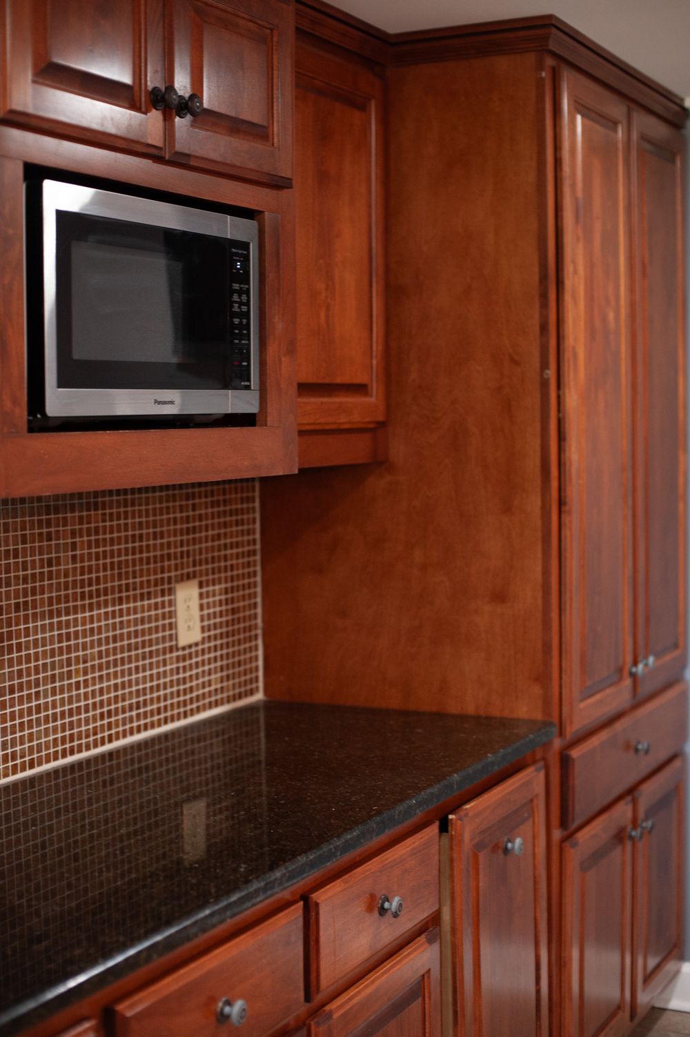 Modern kitchen with dark cherry wood cabinets, black granite countertops, and brown tile backsplash under microwave.