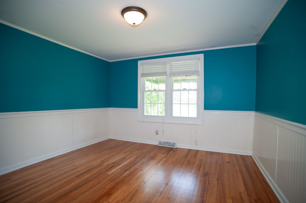 Bedroom with teal walls, white wainscoting, hardwood floors and ceiling fan featuring natural light from window.