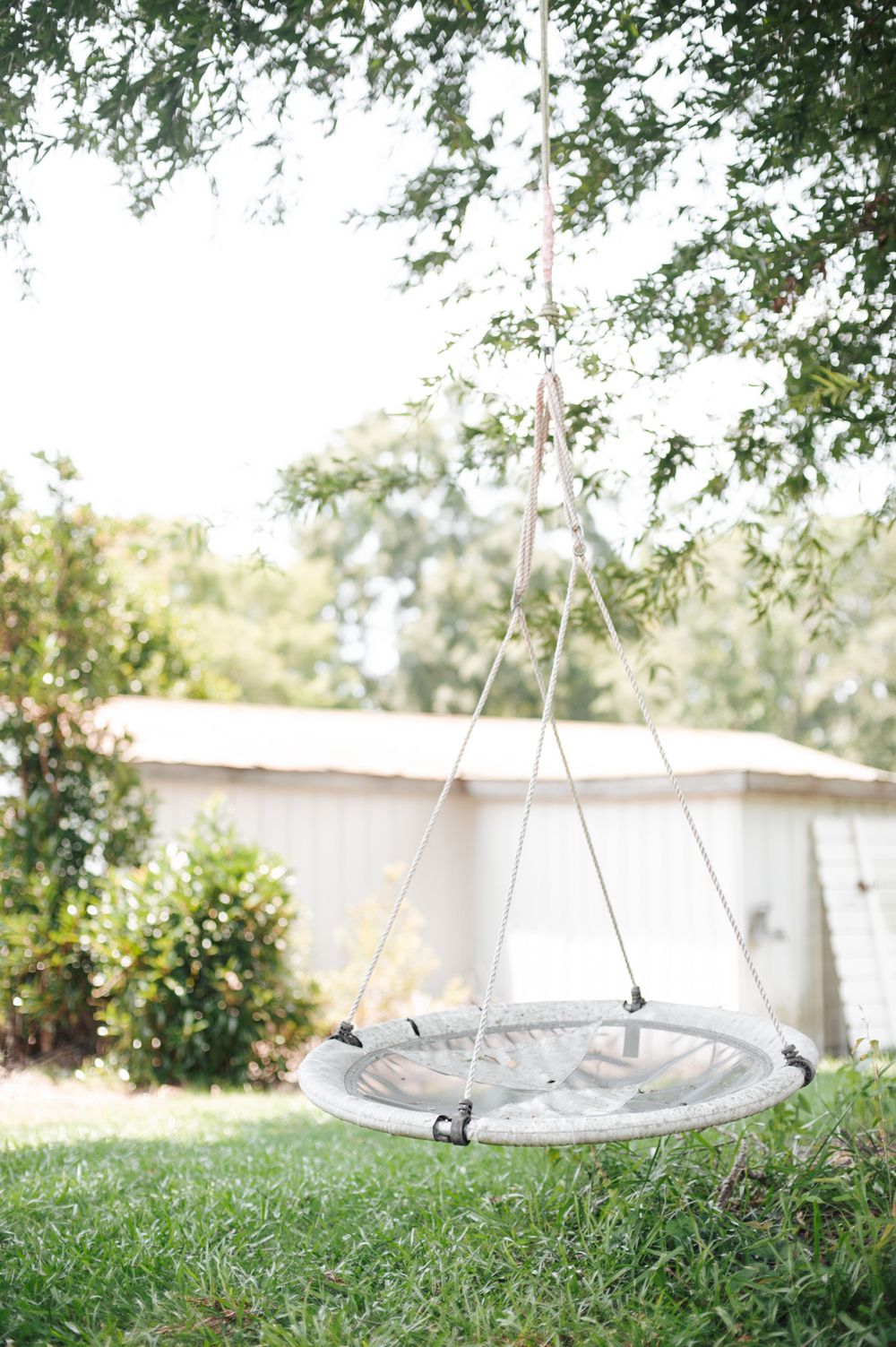 A round gray outdoor swing chair hangs from ropes in a peaceful backyard garden setting with green foliage.