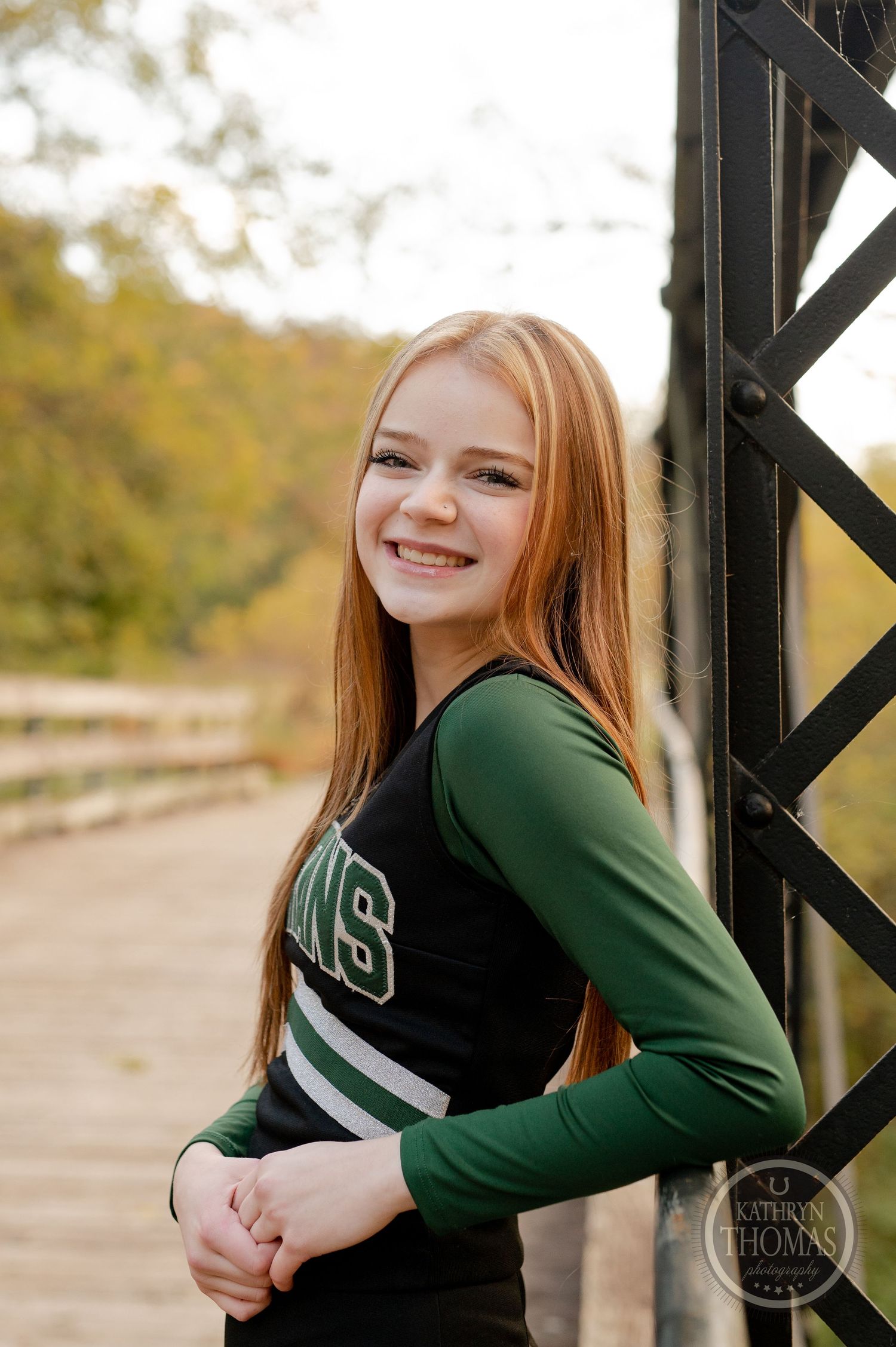 Cheerleader in green and black uniform poses against metal bridge structure during autumn.