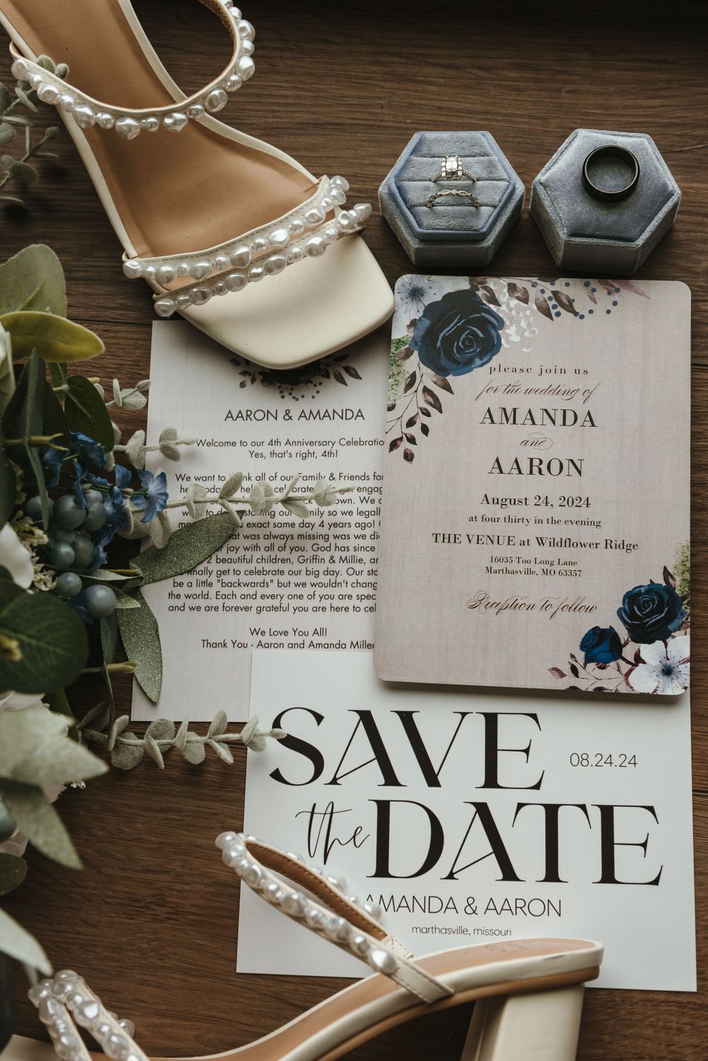 Wedding invitation suite with pearl-adorned shoes and ring boxes on dark wood surface.