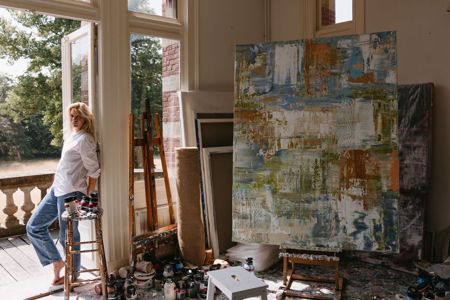 Artist working in sunlit studio with large abstract painting and art supplies.