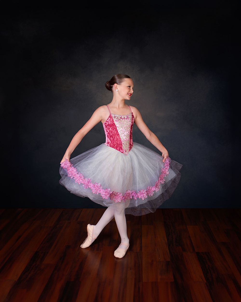 A dancer in a white tutu with pink trim and red sequined bodice poses gracefully on a hardwood floor against dark backdrop.