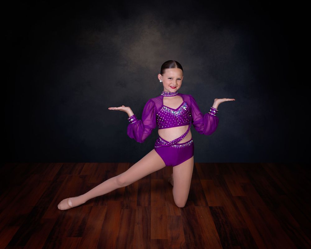 A dancer in a sparkly purple costume strikes a pose on one knee against a dark backdrop on a wooden floor.