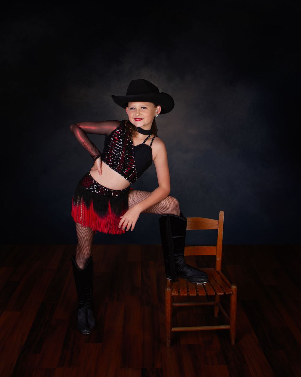 A dancer in red and black outfit performs dramatic poses on a wooden chair against a dark studio backdrop.
