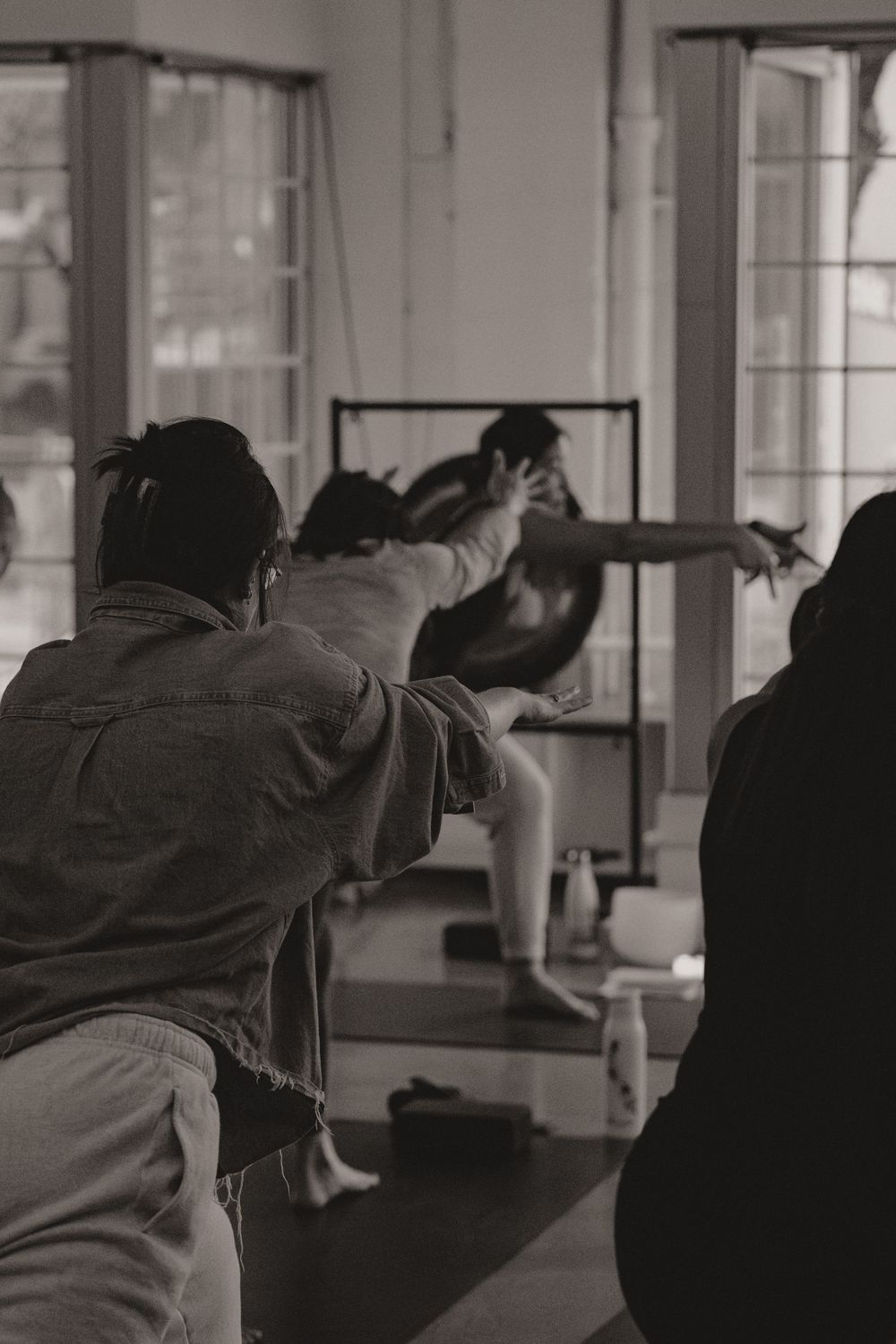 A yoga instructor demonstrates a pose during a class in a sunlit studio with large windows.