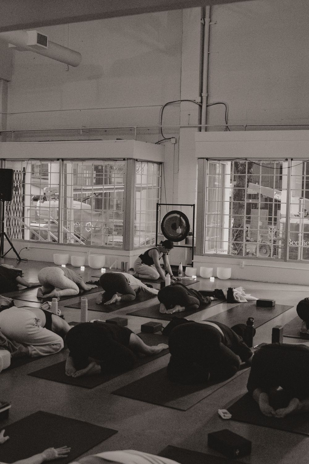 Students practice yoga poses on mats in a bright studio space with industrial windows and exposed ceiling.