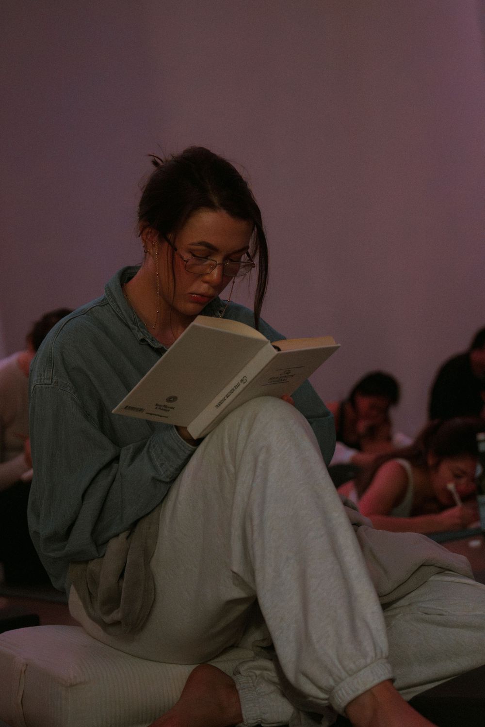 Someone in glasses and casual clothing sits comfortably while reading a book in a dimly lit room.