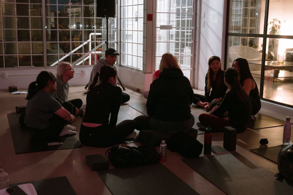 Small group sitting in circle on floor of studio space having discussion in low lighting.