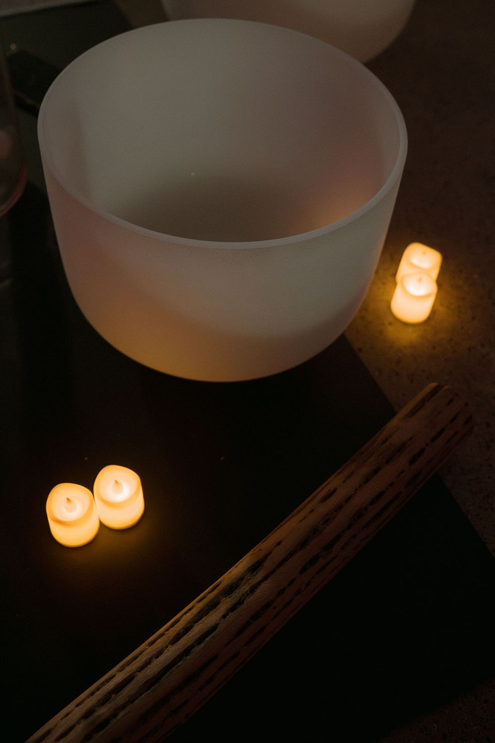 A white crystal singing bowl sits beside glowing candles in a dark meditative setting.