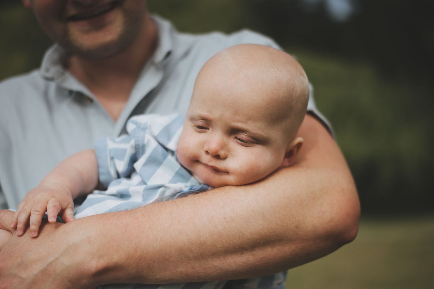 A baby in light blue clothing smiles softly while being held in his father's arms outdoors.