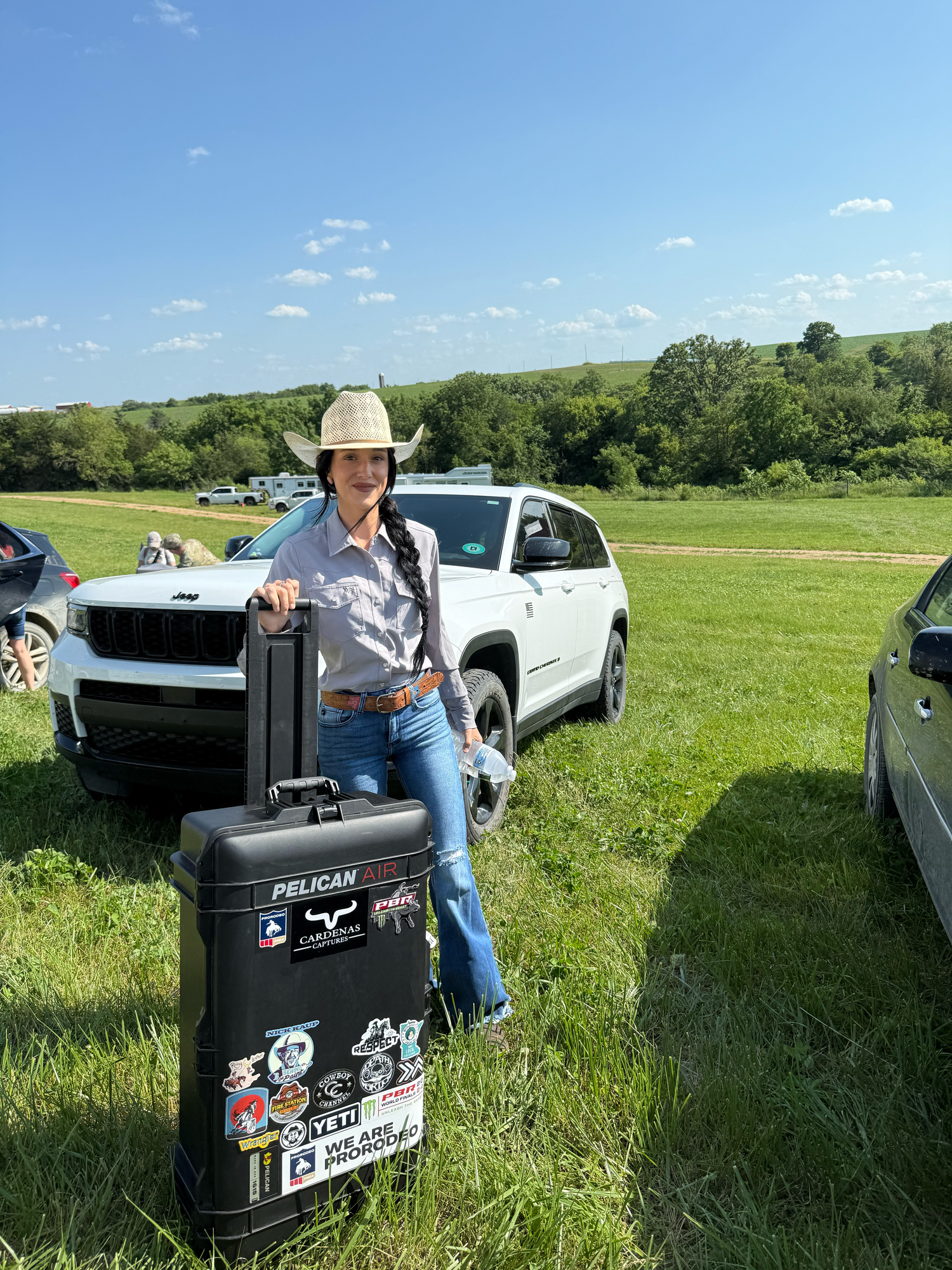 Someone in western attire stands in a grassy field next to a large black equipment case with vehicles parked nearby.