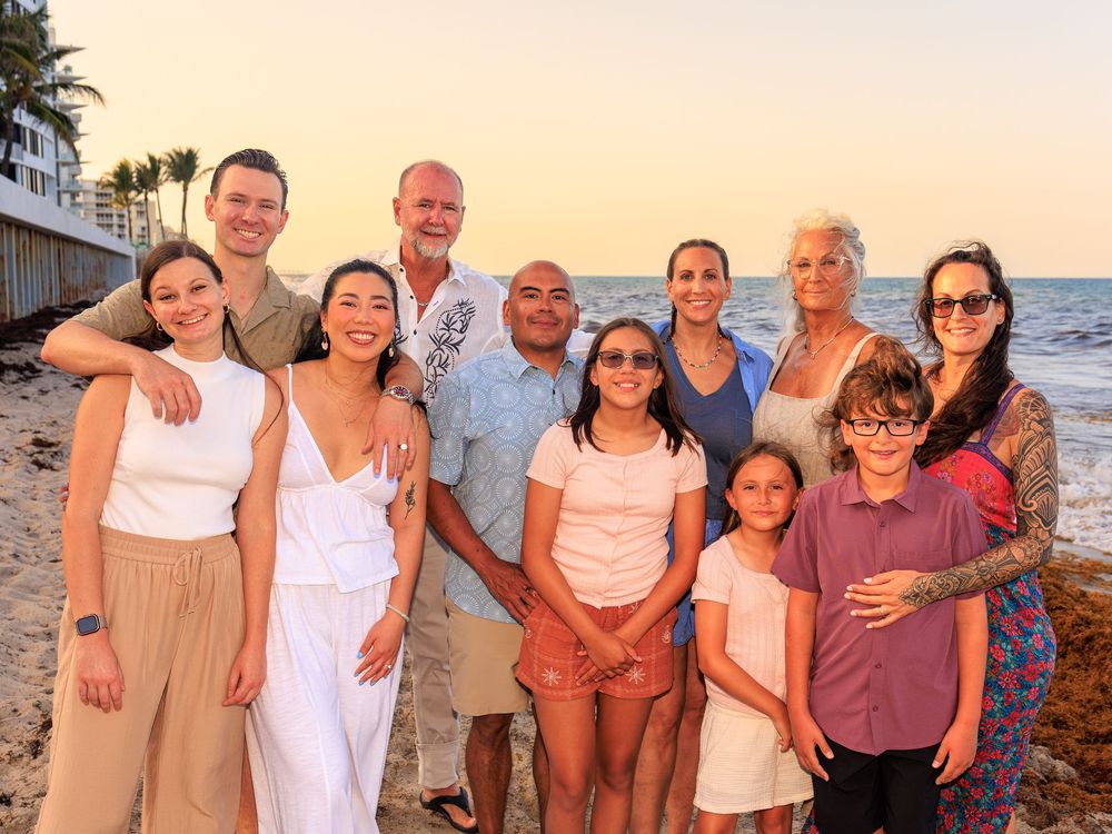 Large family group posing together on a sandy beach at sunset with palm trees and ocean in the background.