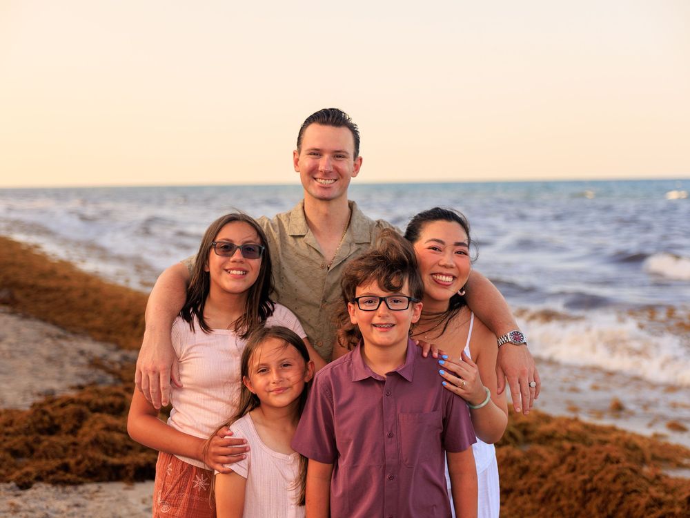 Family of five smiling together on a beach at sunset, with ocean waves in the background.