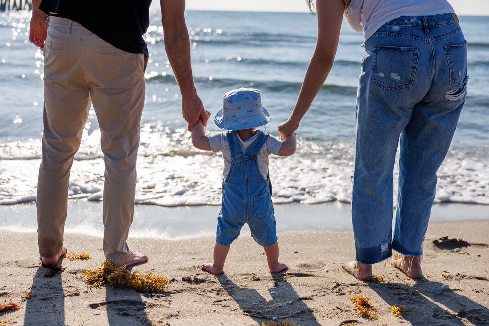 A toddler in blue overalls holds parents hands on a sunny beach with ocean waves in the background.