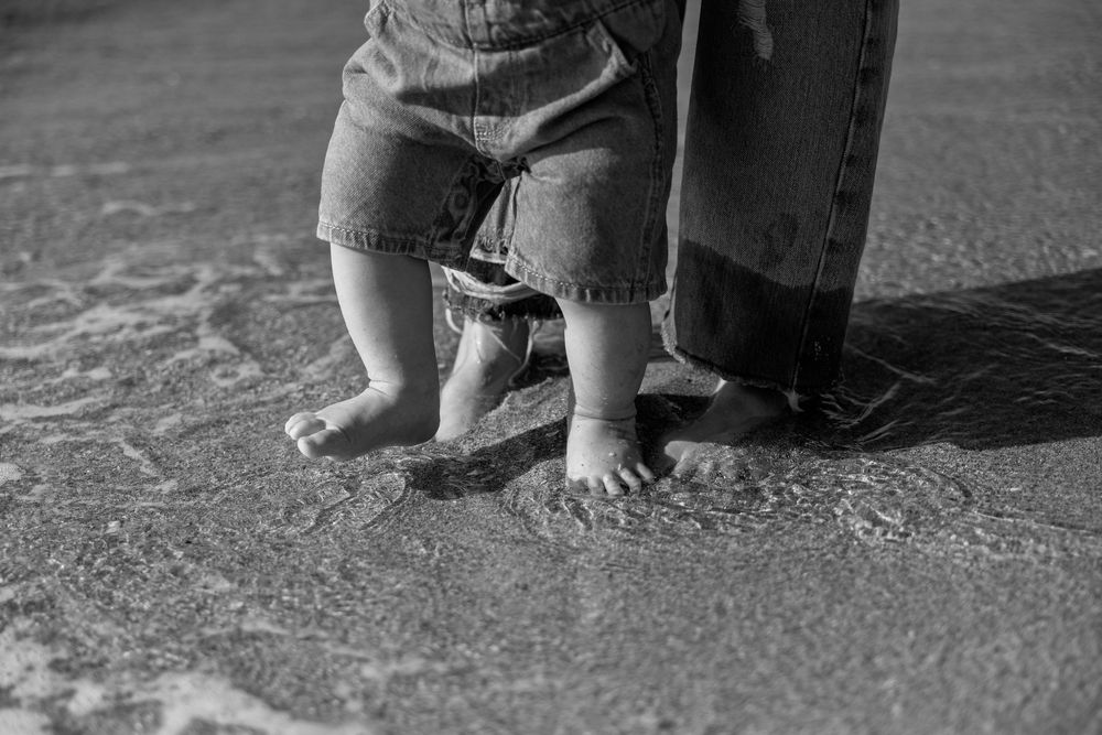 Black and white photo of a baby and adult standing barefoot together on a sandy beach with ocean waves.