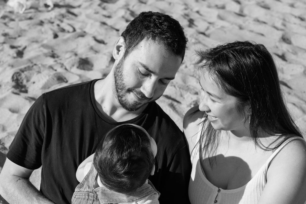 Black and white photo of a couple smiling at their baby on a sandy beach.