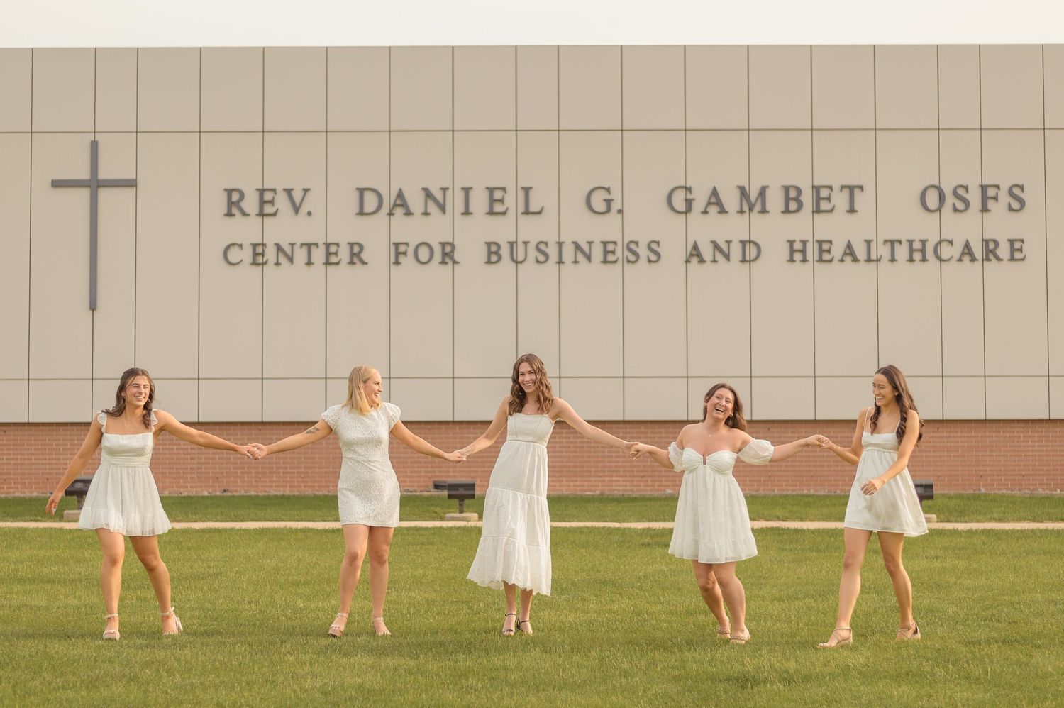 Graduates in white dresses toss their caps in celebration outside university building.