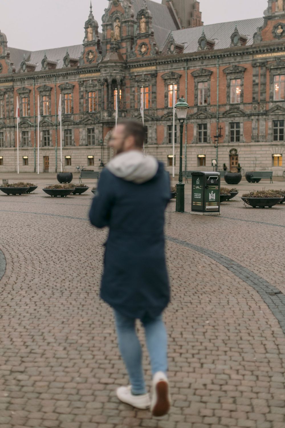 Man in navy blue parka walking across historic cobblestone square with ornate red brick building backdrop.