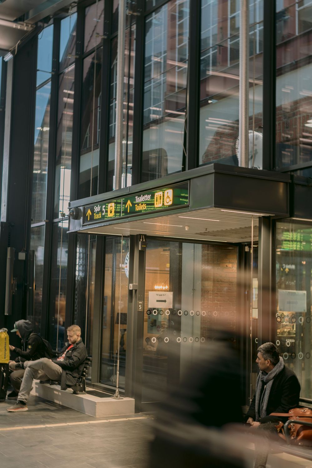 Busy modern transit station entrance with glass facade, directional signs, and commuters waiting outside.