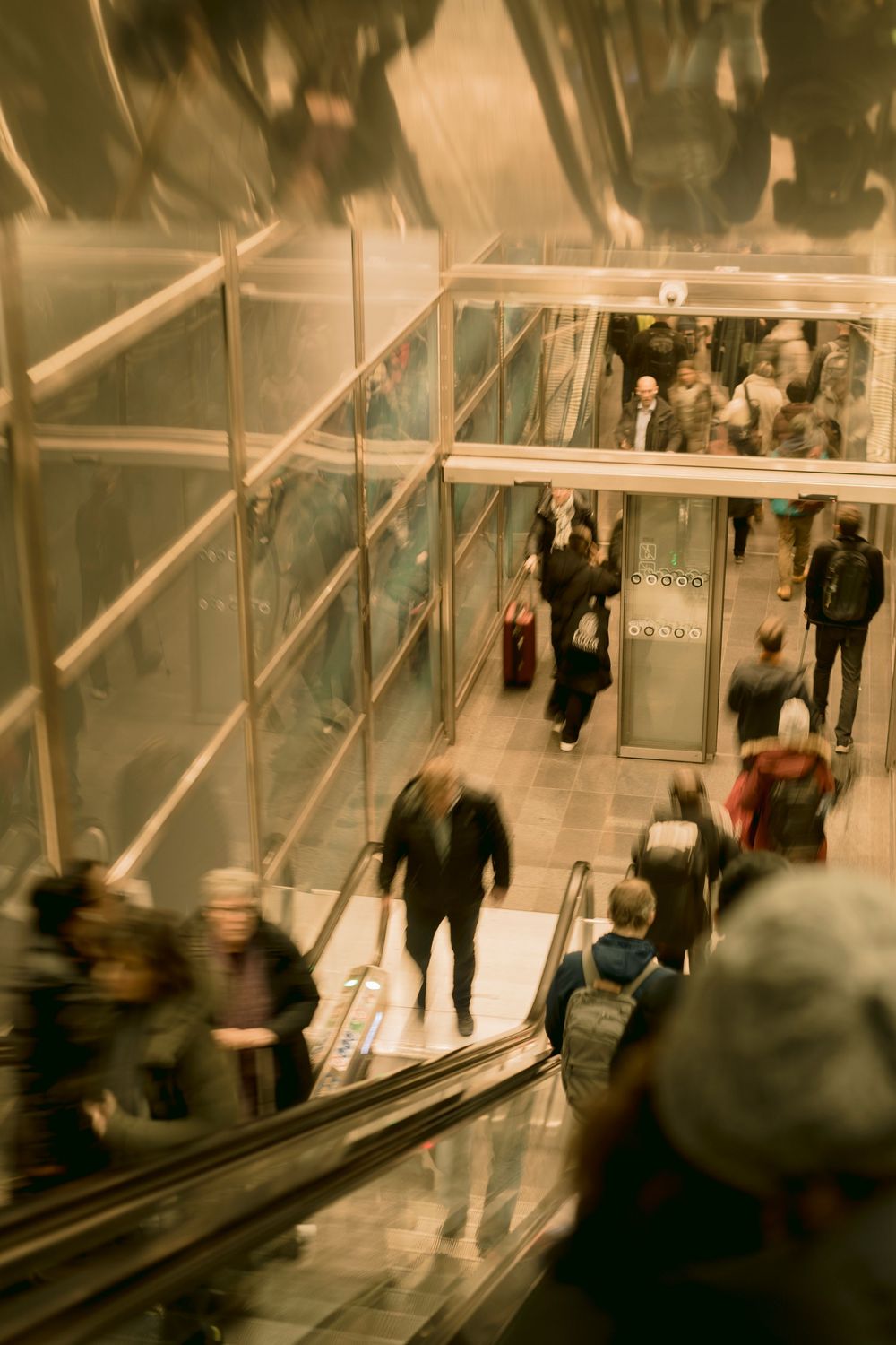 Blurred motion shot of crowds on escalators inside a busy glass-walled shopping center or transit hub.