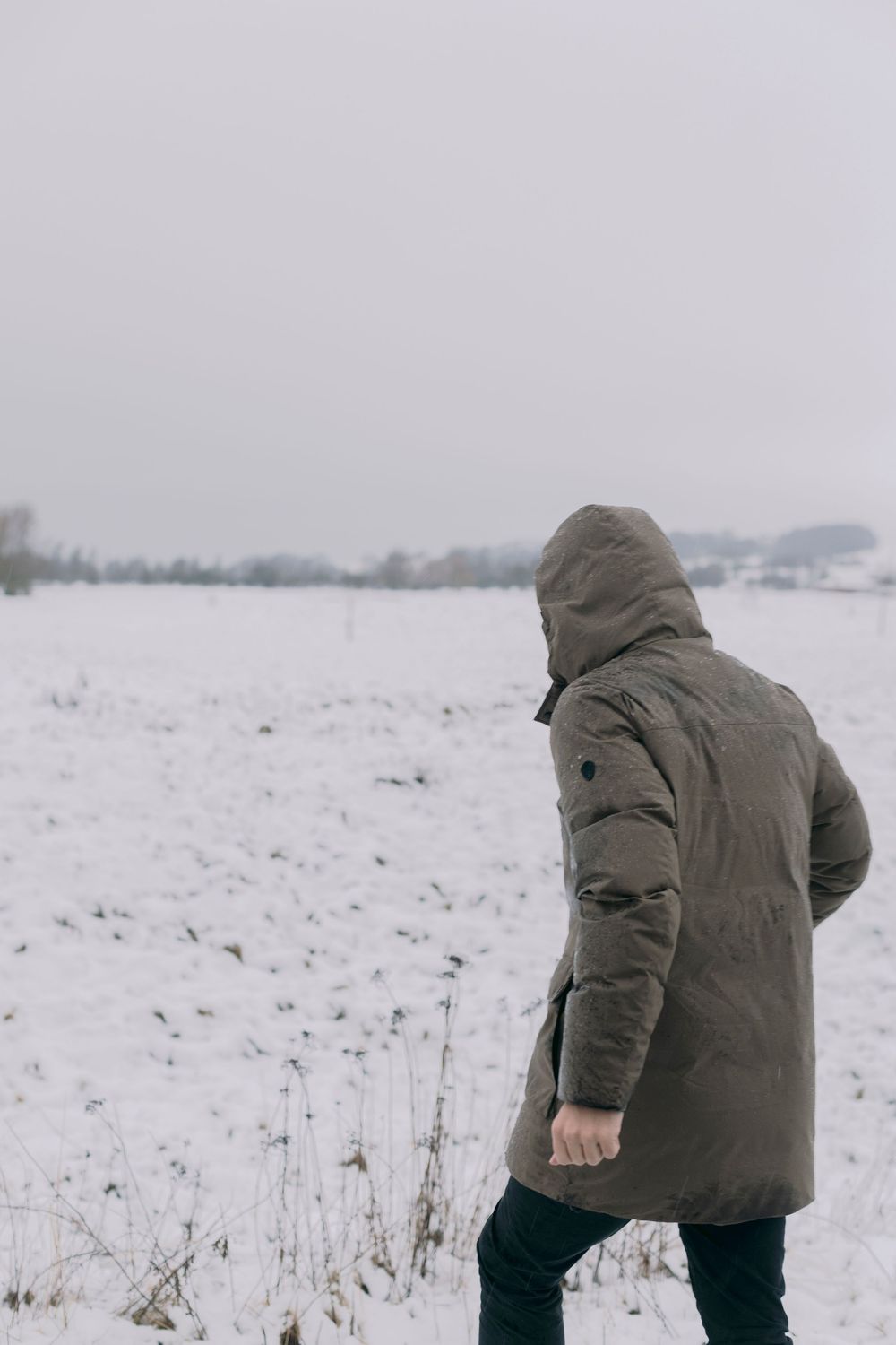Person in olive hooded parka walking through snow-covered winter field under overcast gray sky.