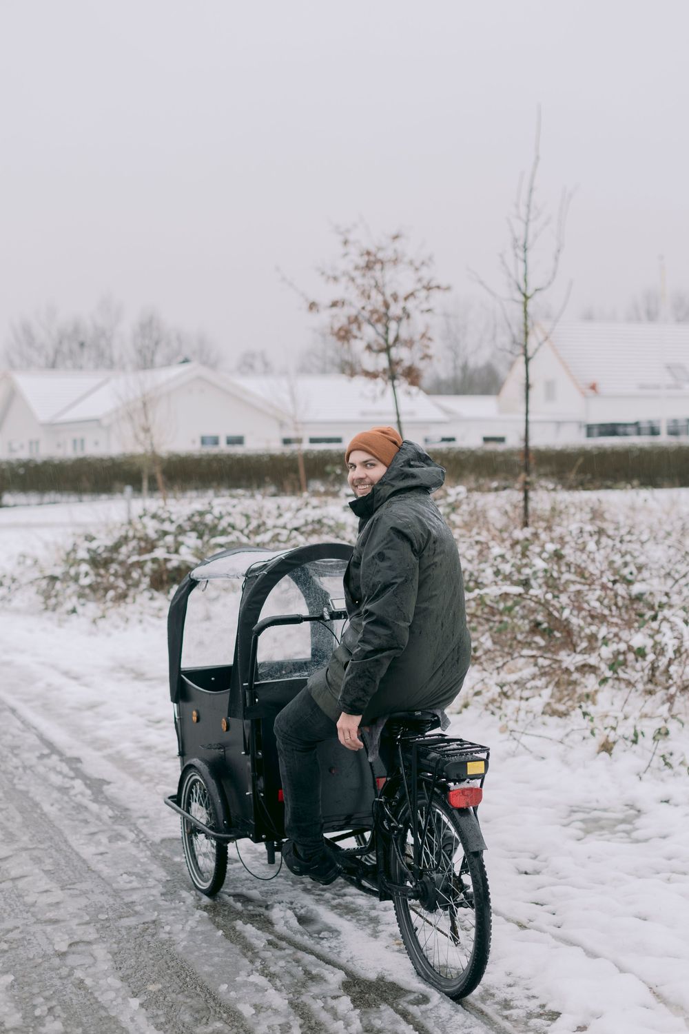 Smiling man riding cargo bike on snowy road in winter with white houses in background.