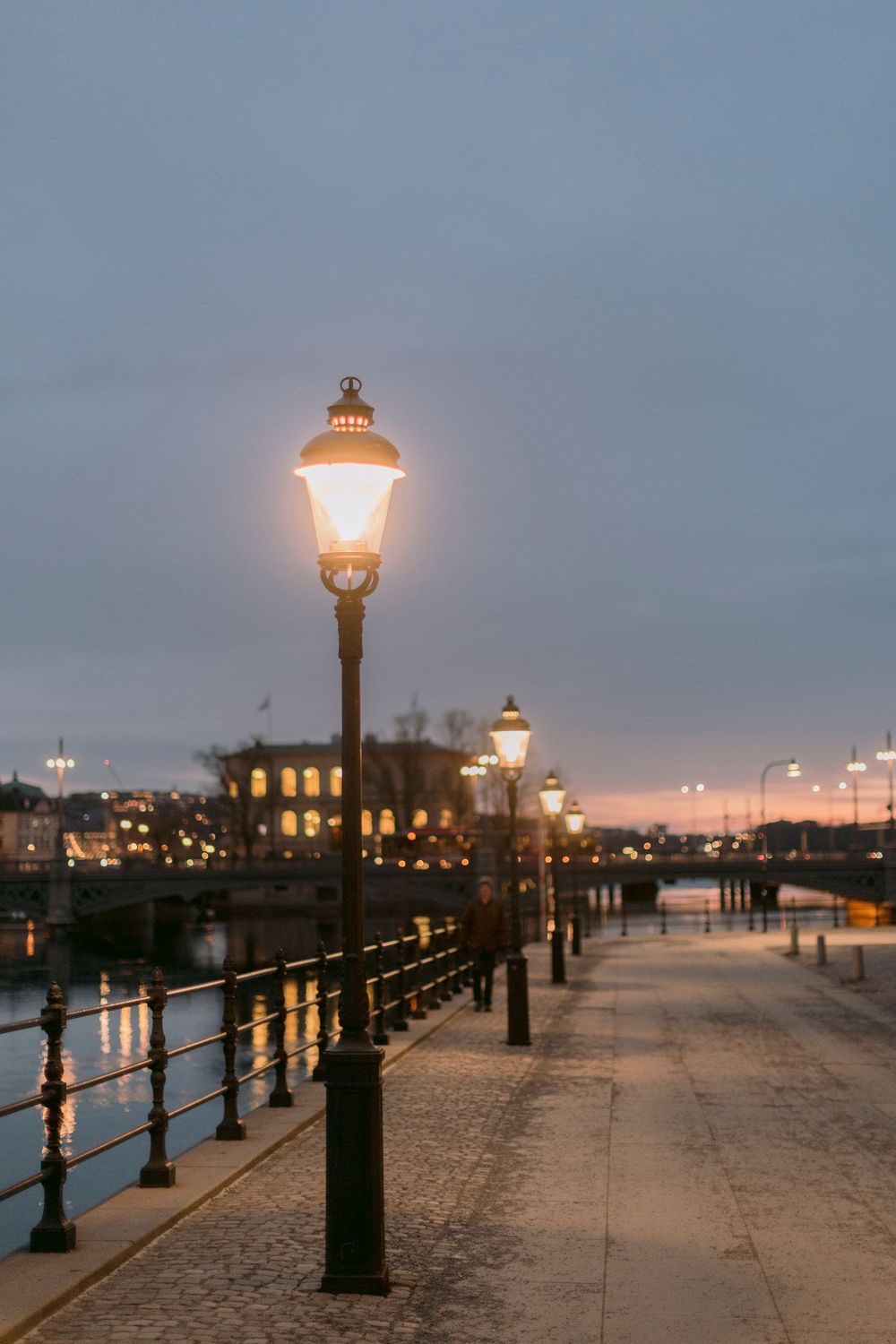 Elegant vintage street lamps glowing along cobblestone waterfront promenade at dusk with city skyline.