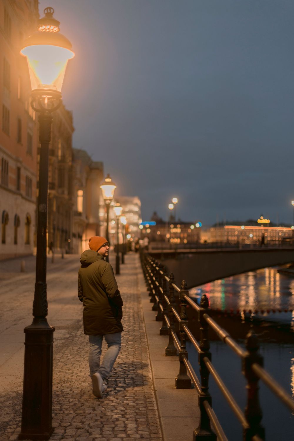 Person walking along illuminated cobblestone waterfront promenade at dusk with glowing street lamps in Stockholm.