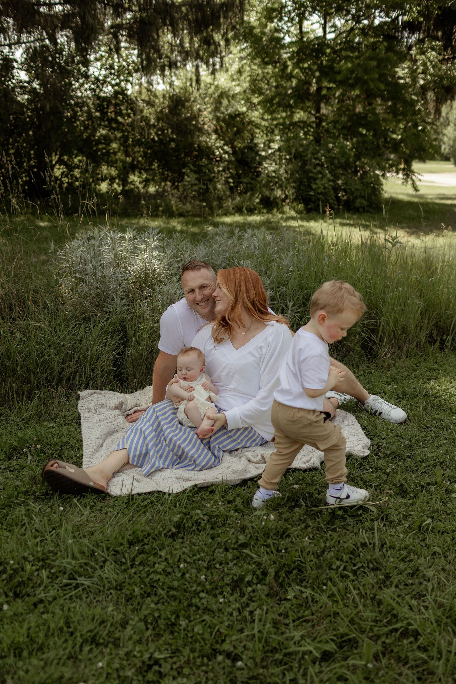 A family sits together on a blanket in a lush green park setting on a sunny summer day.
