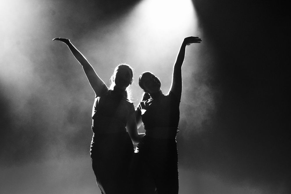 Two silhouettes with raised arms dancing in dramatic backlit fog on stage.