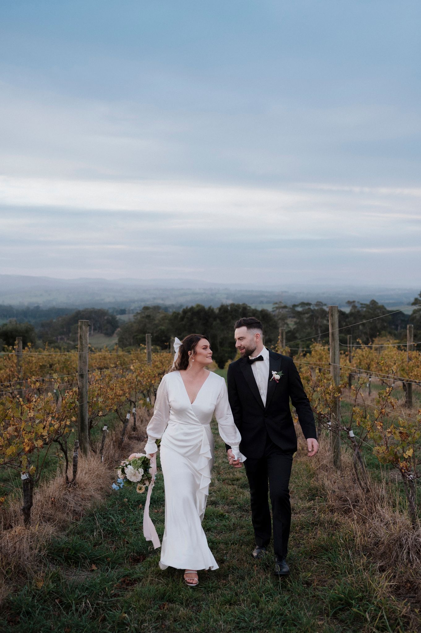 Couple walking through autumn vineyard at sunset wearing formal wedding attire.