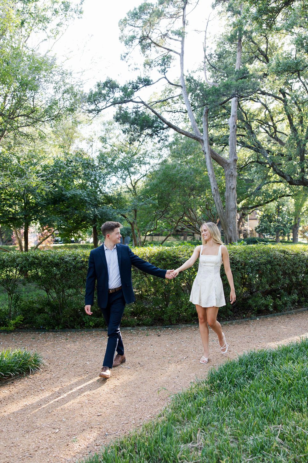 Couple holding hands walking on a garden path, man in navy suit and woman in white mini dress, surrounded by lush greenery.