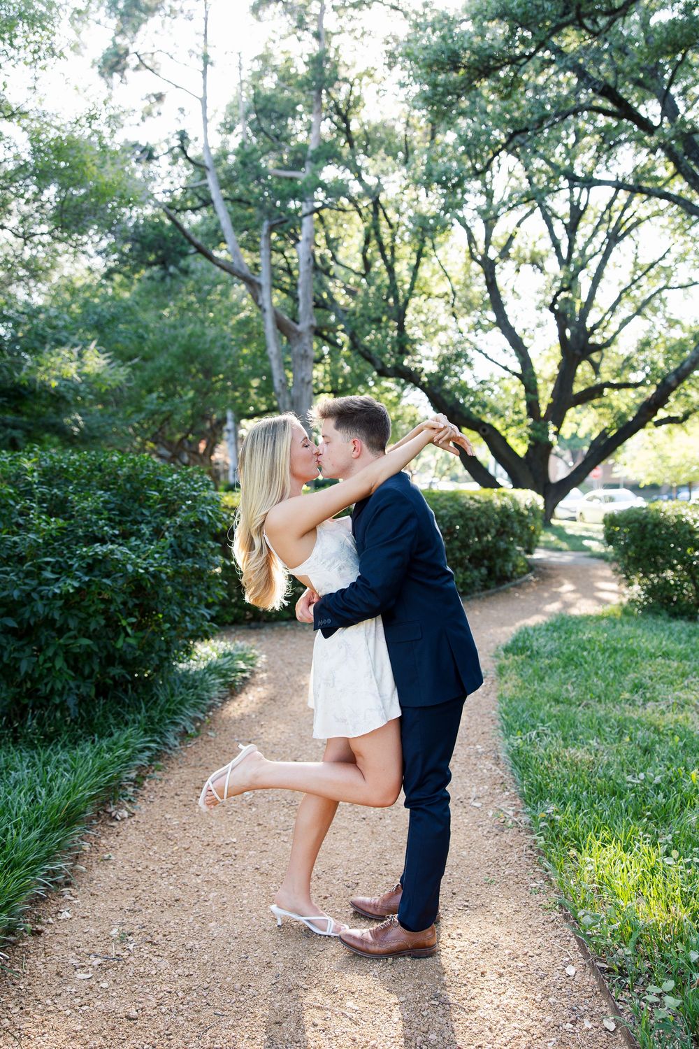 Couple sharing a romantic kiss on a garden path, man in navy suit dipping woman in white dress among green trees.