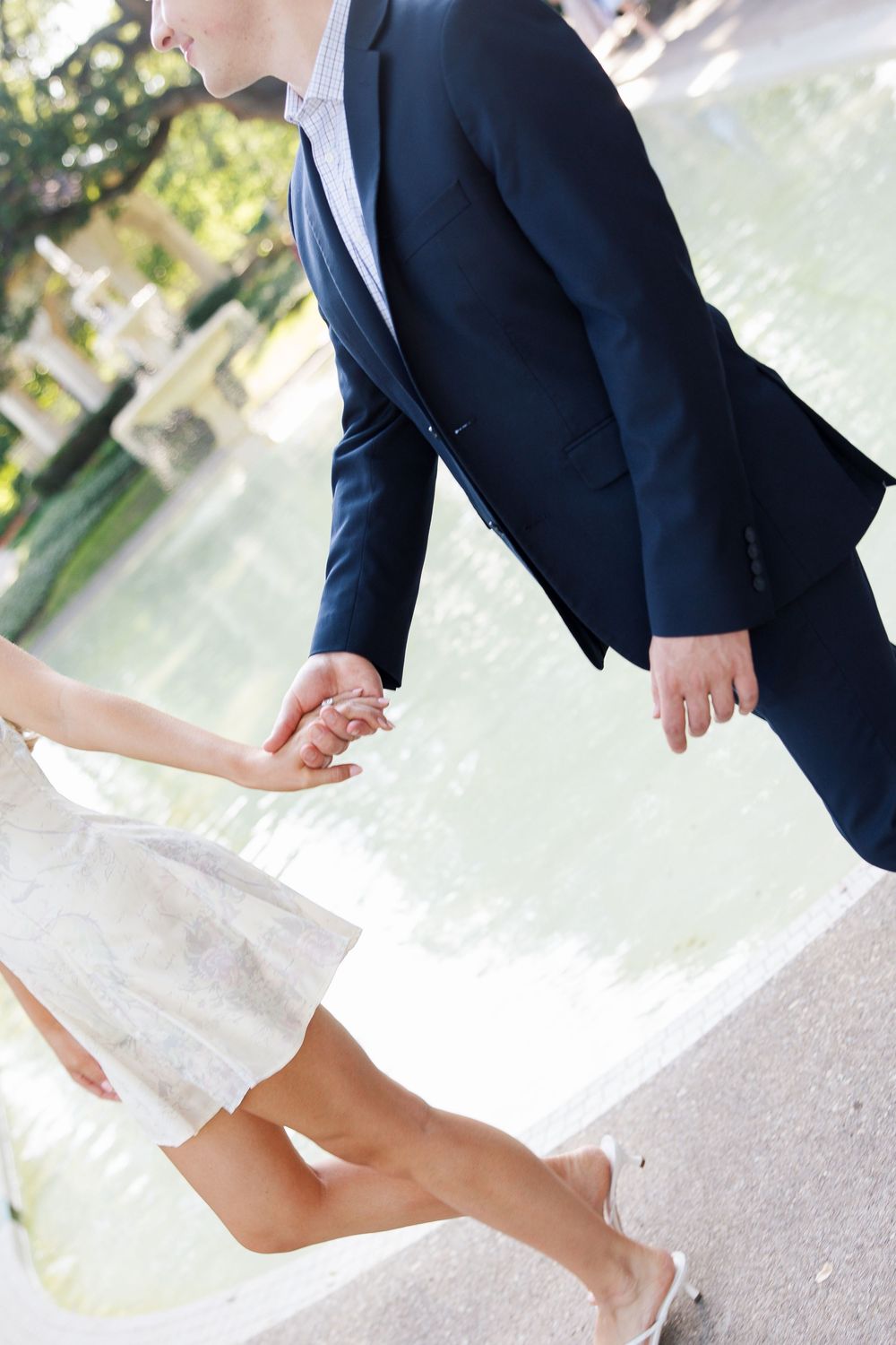 Couple holding hands near a fountain, woman in white dress and man in navy suit, captured from waist down outdoors.