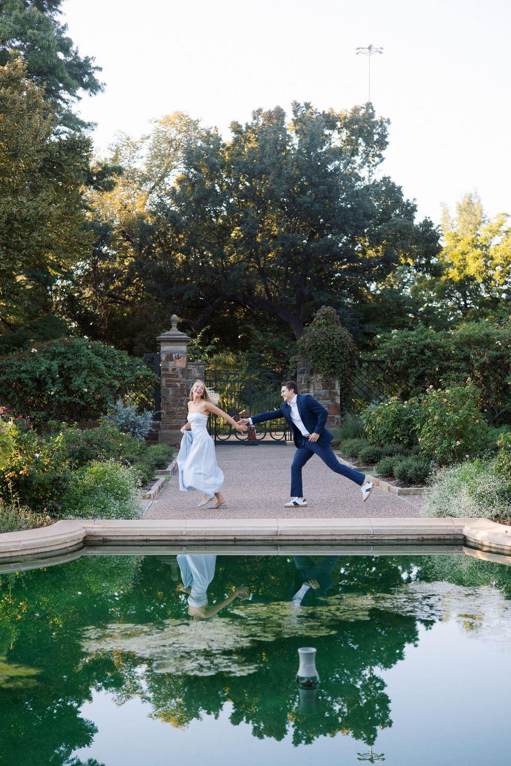 Bride and groom holding hands dancing by a reflective garden pond surrounded by lush greenery and stone gates.