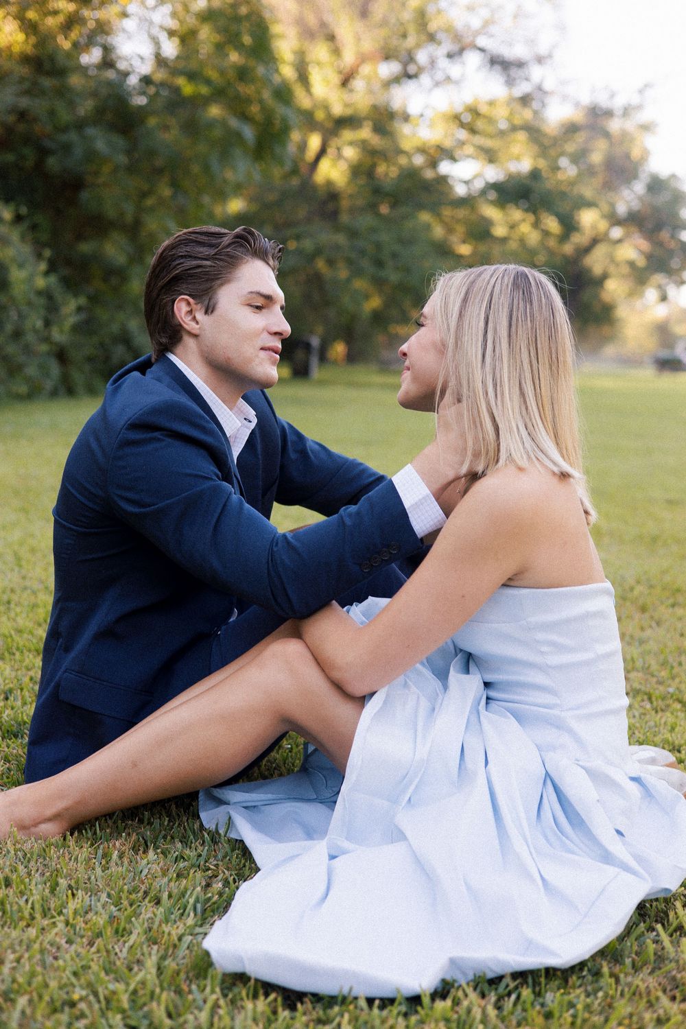Couple sitting on grass in a park, man in navy suit and woman in light blue dress sharing a romantic moment.