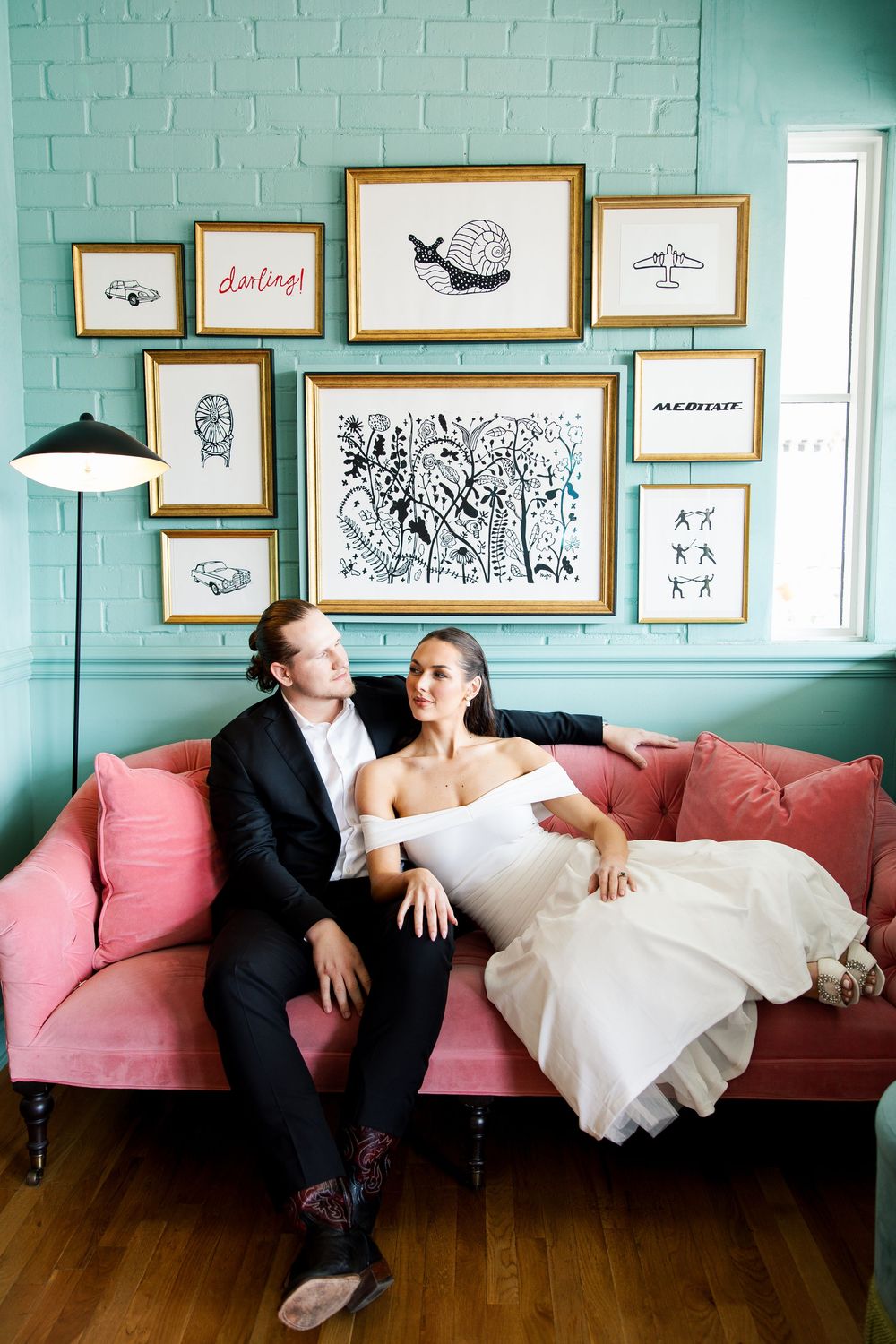 Bride and groom relaxing on a pink velvet sofa beneath a gallery wall in a teal room on their wedding day.