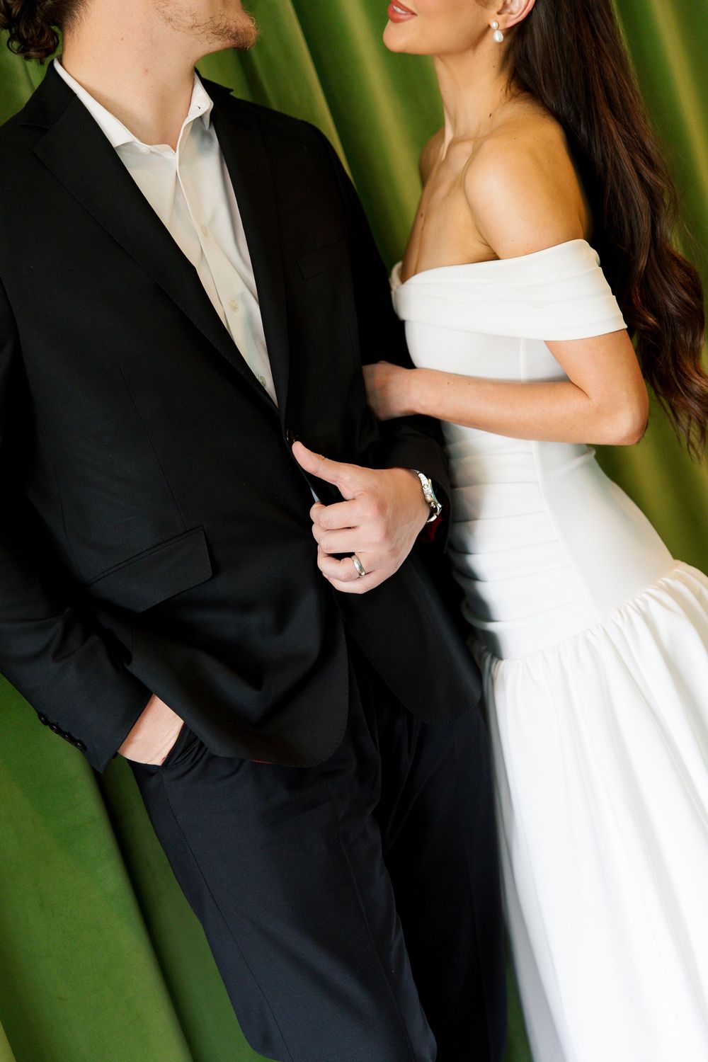 Bride in off-shoulder white gown holds groom's black suit arm against green velvet backdrop.